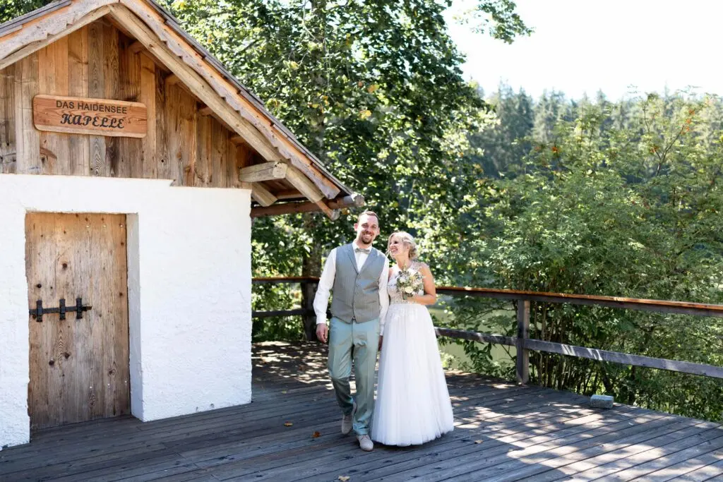 Hochzeit am dasHaidensee – Brautpaar auf dem Steg Brautpaar in Weiß und Anzug geht Hand in Hand über den Steg am dasHaidensee mit Kapelle im Hintergrund