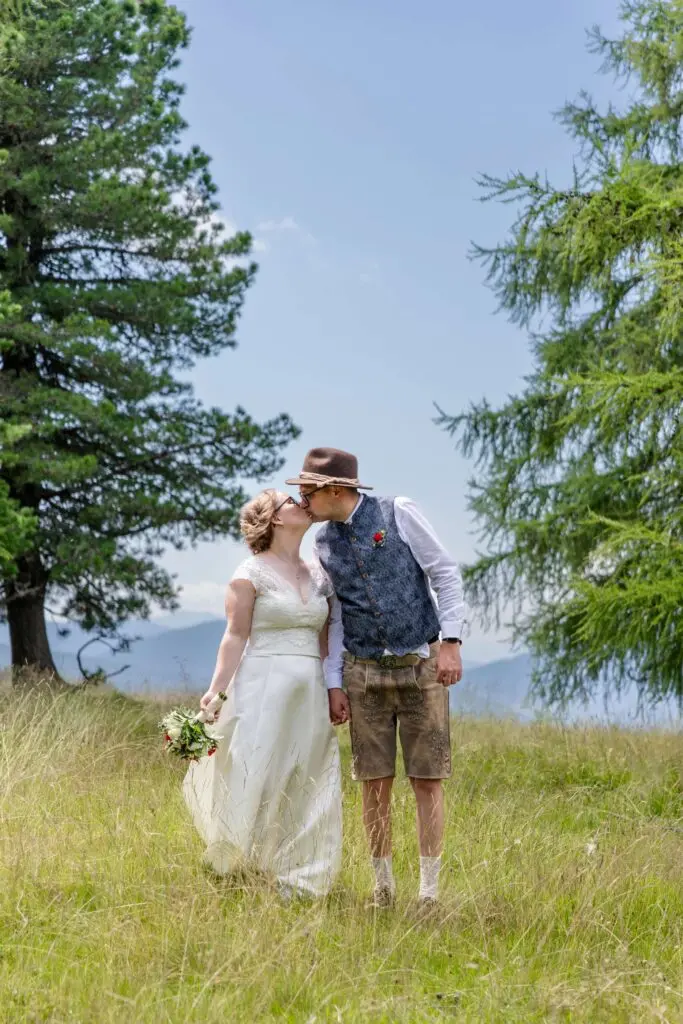 Hochzeit auf der Hochrindl Alm – Brautpaar beim Kuss Braut im weißen Hochzeitskleid und Bräutigam in Tracht küssen sich auf der Hochrindl Alm